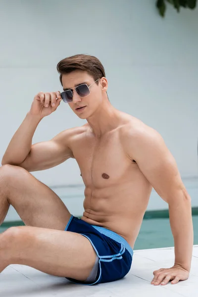 Shirtless man with athletic body adjusting sunglasses while sitting at poolside — Stockfoto