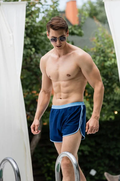 Man with sportive body standing near pool ladder in swimming trunks and sunglasses — Stock Photo