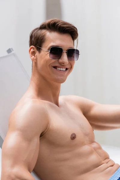 Shirtless man in sunglasses smiling at camera while sitting in deck chair — Photo de stock