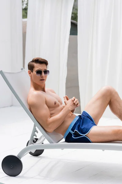 Shirtless man in sunglasses and swimming trunks resting in deck chair at resort — Stockfoto