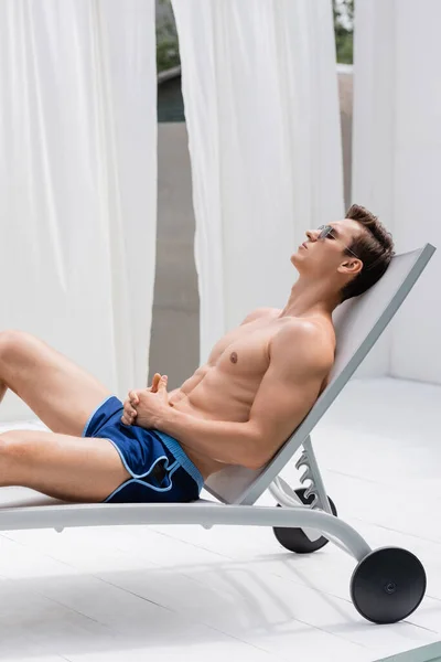 Side view of sportive man in swimming trunks and sunglasses relaxing in deck chair — Stock Photo