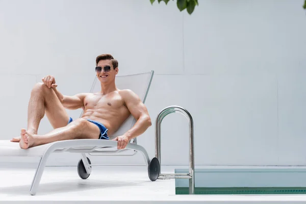 Full length of happy athletic man in sunglasses resting in deck chair near pool — стоковое фото