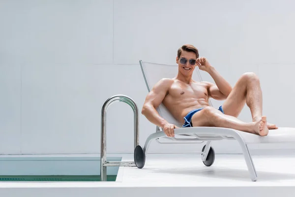 Веселий без сорочки чоловік в сонцезахисних окулярах відпочиває в кріслі на палубі біля басейну — стокове фото