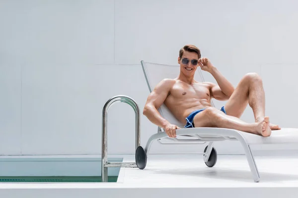 Cheerful shirtless man in sunglasses resting in deck chair near pool - foto de stock