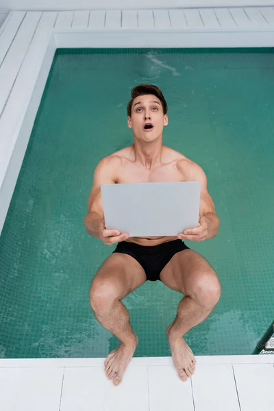 Vista dall'alto dell'uomo entusiasta con la bocca aperta che cade in piscina con il computer portatile — Foto stock