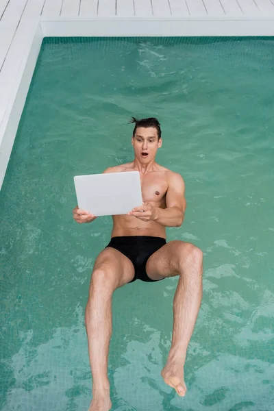 Top view of excited man with open mouth falling in pool with laptop — Stock Photo