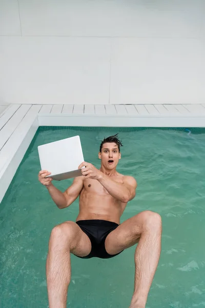 Изумленный человек падает в бассейн с ноутбуком, глядя в камеру — стоковое фото