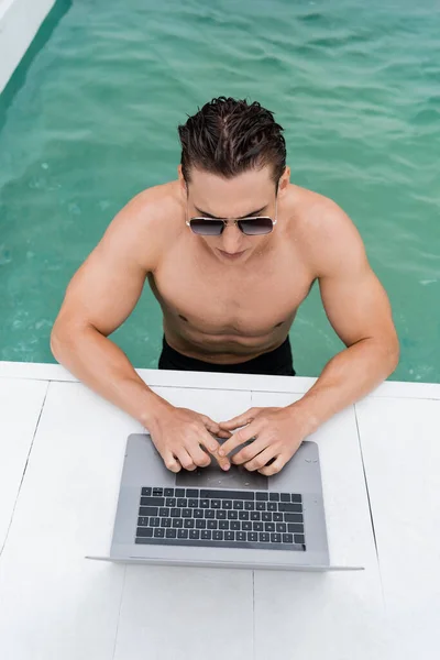 Мокрий і спортивний чоловік в сонцезахисних окулярах використовує ноутбук біля басейну — стокове фото
