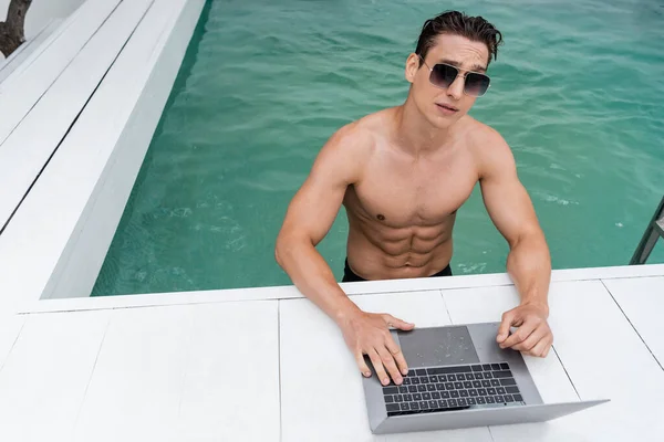 Sportlicher Mann mit Sonnenbrille mit Laptop am Pool — Stockfoto