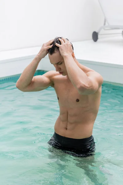 Man with muscular body standing in pool and touching wet hair — стоковое фото