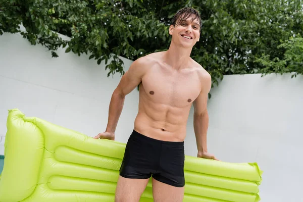 Happy man in swimming trunks holding inflatable mattress and looking at camera - foto de stock