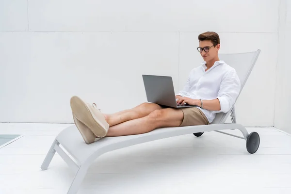 Full length of smiling freelancer in summer clothes sitting in deck chair and typing on laptop — Stockfoto