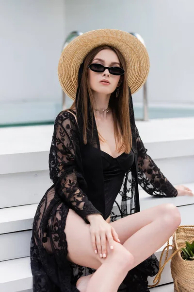 Stylish young woman in swimsuit and sunglasses sitting near handbag on stairs at resort — Photo de stock