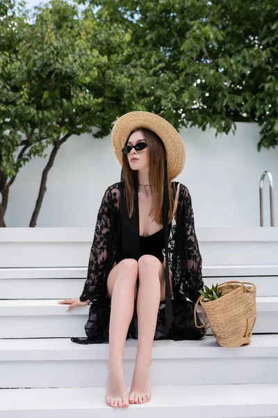 Fashionable woman in guipure robe and sunglasses sitting near handbag on stairs at resort — Photo de stock