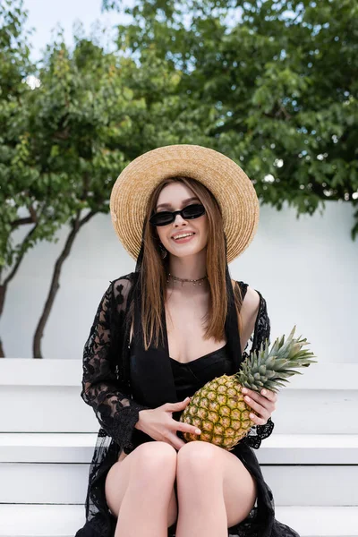 Cheerful woman in straw hat and swimsuit holding pineapple at resort — Stockfoto