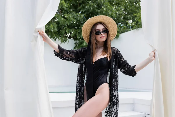 Fashionable woman in swimsuit and sunglasses holding curtains at resort — Photo de stock