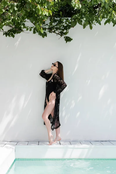 Stylish woman in guipure robe and swimsuit standing near wall and swimming pool — Foto stock