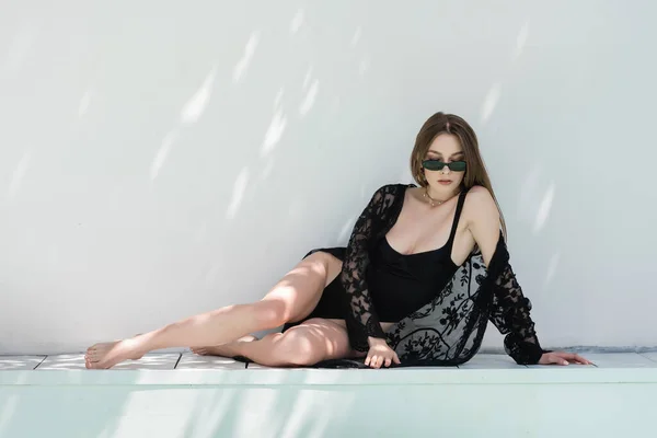 Stylish young woman in robe and swimsuit sitting near wall with sunlight at resort — Stock Photo