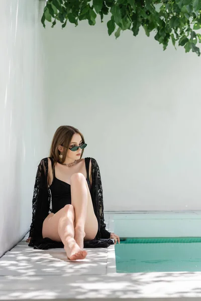 Trendy woman in sunglasses and swimsuit looking away near swimming pool at resort — стоковое фото