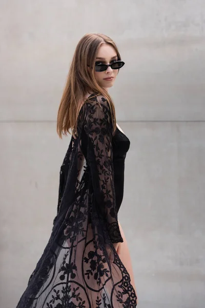 Pretty young woman in sunglasses and black swimwear looking at camera at resort — Stock Photo