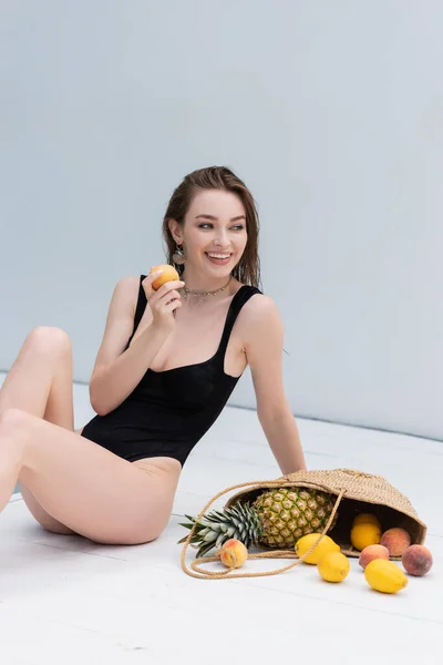 Cheerful young woman in swimsuit holding peach near fruits in straw handbag at resort — Fotografia de Stock