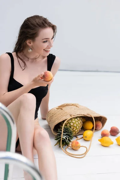 Smiling woman in swimwear holding fresh peach near fruits in straw handbag at resort — Foto stock
