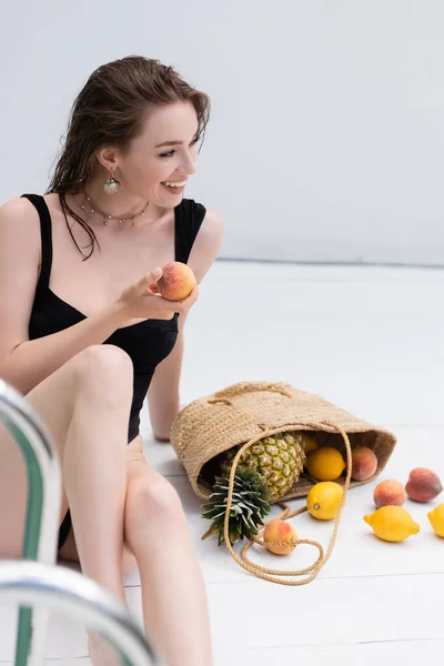 Smiling woman in swimwear holding fresh peach near fruits in straw handbag at resort — стоковое фото