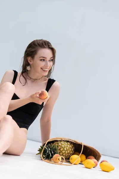 Cheerful woman in swimwear holding peach near tropical fruits in handbag at resort — Foto stock