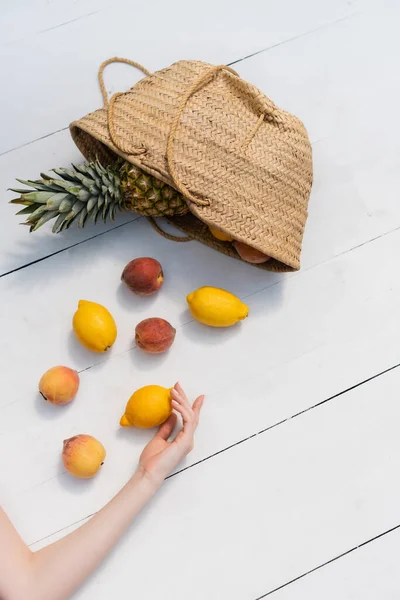 Top view of woman touching fresh lemon near fruits in straw handbag on floor — стоковое фото