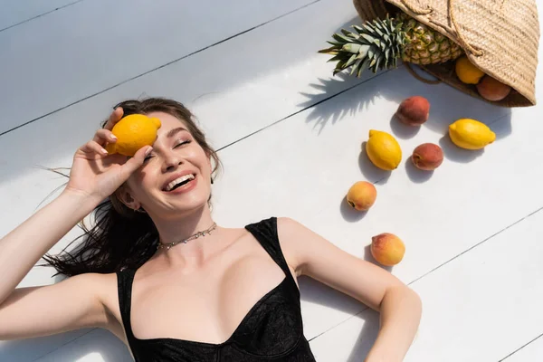Vista superior da jovem mulher feliz em maiô preto deitado perto de frutas frescas na superfície branca — Fotografia de Stock