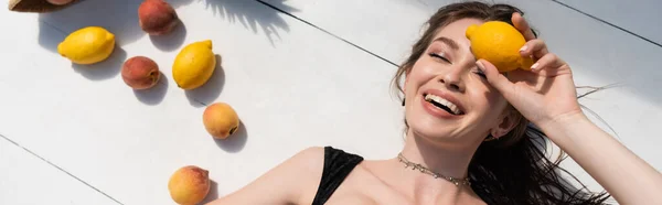 Top view of happy young woman in black swimsuit lying near fresh fruits on white surface, banner — Foto stock