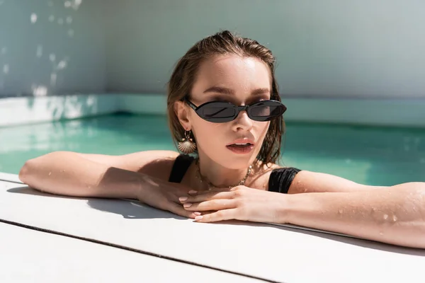 Portrait of stylish woman in black sunglasses and golden accessories resting in swimming pool — стоковое фото