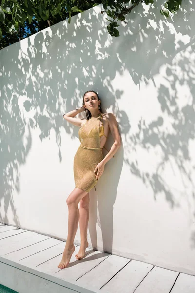 Full length of woman in golden summer dress with swimwear posing near shadows on wall and pool — Stock Photo