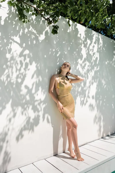 Comprimento total de descalça jovem mulher em vestido de verão dourado de pé perto enquanto parede com sombras de folhas verdes — Fotografia de Stock