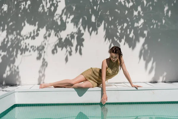 Стильна жінка в літньому платті і купальнику позує біля басейну і торкається води — стокове фото