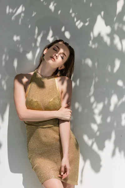 Красива молода жінка з закритими очима позує в літній сукні біля білої стіни з тінями від листя — стокове фото