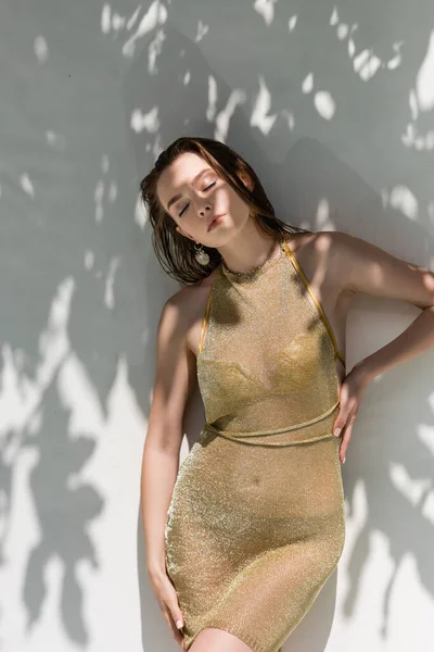 Pretty young woman with closed eyes posing with hand on hip near white wall with shadows from leaves — Fotografia de Stock