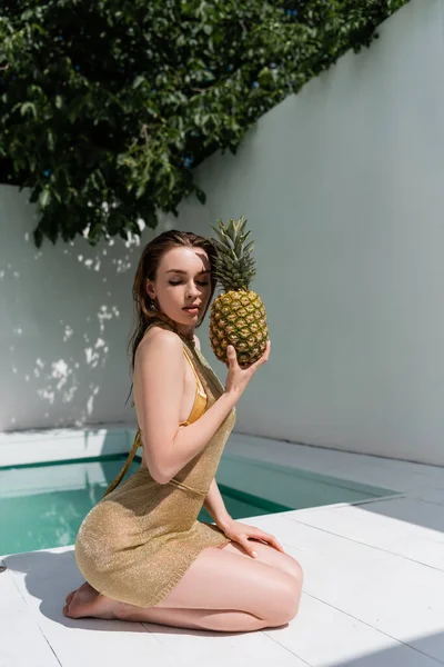 Pretty young woman in golden summer dress sitting with fresh pineapple at poolside — Foto stock