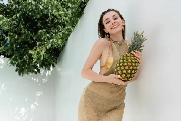 Happy young woman in golden summer dress holding ripe pineapple near white wall — Photo de stock