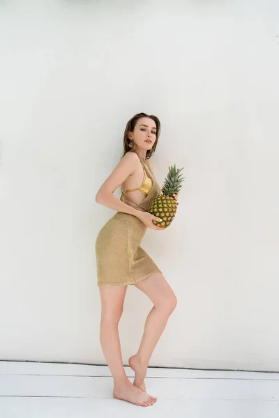 Full length of young woman in golden summer dress holding fresh pineapple near white wall — Stock Photo