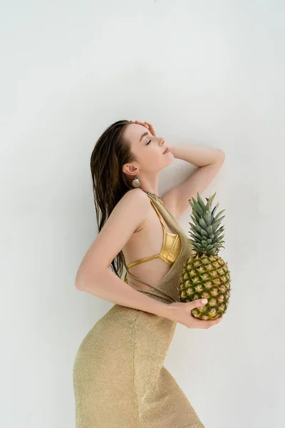 Side view of smiling woman in golden swimwear and dress holding ripe pineapple near while wall - foto de stock
