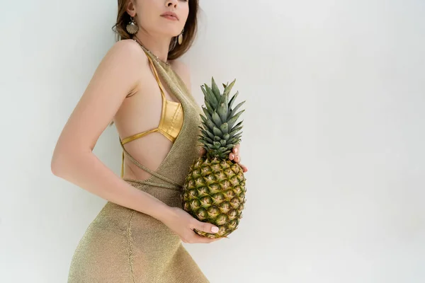 Cropped view of woman in golden swimwear and dress holding ripe pineapple near while wall — Stockfoto