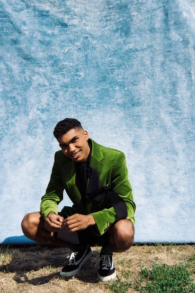 Smiling african american man in green velvet blazer sitting on haunches near blue cloth — Foto stock