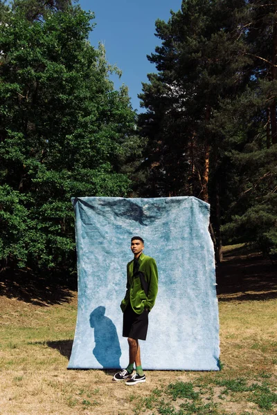 African american man in green blazer posing with hands in pockets of black shorts near blue cloth in park — Stock Photo