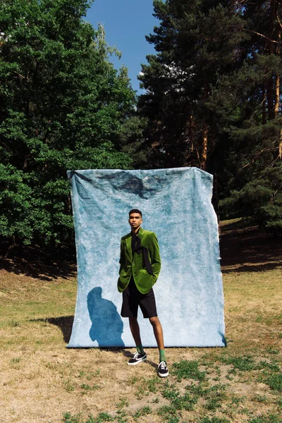 Full length of african american man in green blazer and sneakers near blue drape in forest — Stock Photo