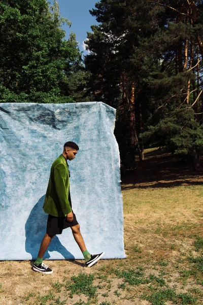 Side view of trendy african american man walking near blue drape in park — стоковое фото