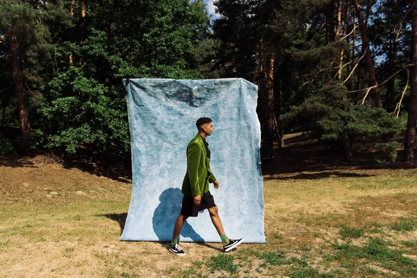 Side view of african american man in green blazer and black shorts walking near blue cloth in park — стоковое фото