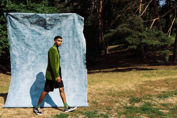 Full length of african american man in velvet blazer and shorts walking near blue cloth in park - foto de stock