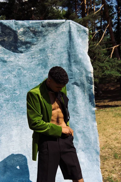 African american man in velvet blazer adjusting black shorts near blue cloth in park — Photo de stock