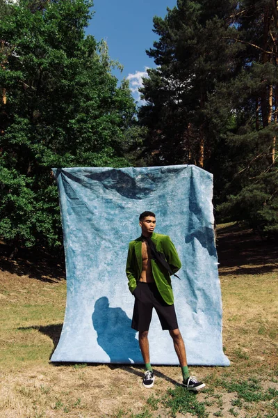 Full length of african american man in sneakers and velvet blazer standing near blue cloth in forest — стоковое фото